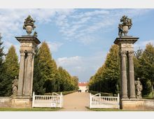 Steinerne Eingangstore mit verzierten Säulen, flankiert von Statuen, führen auf einen breiten Kiesweg, der von Bäumen gesäumt ist. Im Hintergrund ist ein helles Gebäude mit rotem Dach sichtbar, eingebettet in die parkähnliche Landschaft.