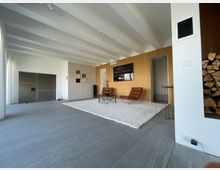 Modernes Wohnzimmer mit großen grauen Bodenfliesen, einer weißen Decke mit freiliegenden Balken und einer Wandverkleidung aus Holz. Im Raum stehen zwei braune Sessel auf einem weißen Teppich, daneben ein kleiner Metalltisch; im Hintergrund befindet sich ein Kaminholzfach und eine noch unfertige Wand.