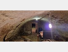 Gewölbekeller mit gemauerten Wänden und einem gewölbten Ziegelsteinbogen an der Decke. Im Raum befinden sich diverse Gegenstände, darunter eine Lampe an einem Kabel, ein Tisch und ein schwarzes Fass, sowie eine geöffnete Tür, die in einen weiteren Bereich führt.