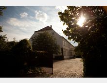 Herrenhaus mit heller Fassade und roten Dachziegeln, umgeben von Bäumen und einem offenen Tor mit Bewuchs. Ein gepflasterter Hof führt zum Gebäude, im Hintergrund strahlt die Sonne durch das Blätterdach.