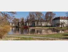 Ein kleiner See in einer parkähnlichen Umgebung, umgeben von Grasflächen, kahlen Bäumen und mehrstöckigen Wohnhäusern im Hintergrund. Ein schmaler Weg führt entlang des Ufers, und trockenes Schilf säumt den Rand des Wassers.