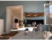 Wohnzimmer mit blauen Wänden, einem weißen Tisch im Vordergrund, darauf mehrere dekorative Gläser und Kerzen. Im Hintergrund befindet sich eine geöffnete Tür mit Blick in einen weiteren Raum, in dem ein Fernseher sowie weiße Möbel stehen; an der Wand hängt ein Holzrelief mit Pferdemotiven und ein Fenster lässt Tageslicht herein.