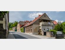 Eine ruhige Dorfstraße mit alten Wohnhäusern, die zumeist aus verputztem Mauerwerk mit roten Ziegeldächern bestehen. Auf der rechten Seite befindet sich ein Gebäude mit einem kleinen Balkon und einem Vorgarten, im Hintergrund sind Bäume und weitere Gebäude sichtbar.