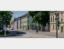 Straßenszene in einer kleinen Stadt mit mehrstöckigen Wohnhäusern, darunter ein pastellgrünes Gebäude und ein cremefarbenes historische Gebäude mit verzierten Fensterrahmen. Eine Ampel zeigt grünes Signal für Fußgänger und ein Auto fährt auf der leicht kurvigen Straße entlang; im Hintergrund Bäume und weitere Gebäude.