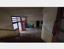 Treppenhaus in einem schlichten Gebäude mit hellgelb gefliesten Wänden und einem Boden aus beigen Fliesen. Unten führen rote Türen aus Holz mit Glaselementen in einen weiteren Raum, in dem ein türkisfarbener runder Empfangstresen steht.