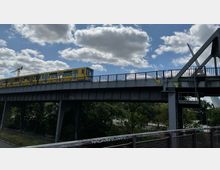 Gelbe U-Bahn fährt auf einer erhöhten Stahlbrücke, darunter Bäume und eine Straße; im Hintergrund blauer Himmel mit vereinzelten Wolken und ein Baukran sichtbar.