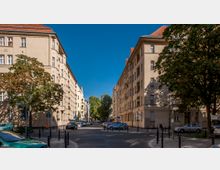 Straßenszene in einer Wohngegend mit mehrstöckigen, beigefarbenen Altbauten mit roten Dächern auf beiden Seiten. Bäume säumen die Straße, geparkte Autos und Fahrräder sind zu sehen, und die Straße ist durch mehrere Straßenpfosten begrenzt.