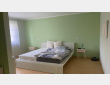 Schlafzimmer mit hellgrünen Wänden, einem Doppelbett mit hellem Kopfteil und gemusterter Bettwäsche. Auf beiden Seiten des Bettes stehen kleine Nachttische, der Boden ist aus Holz und links sind bodenlange weiße Vorhänge an einem Fenster.