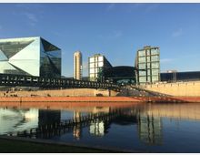 Modernes Stadtbild mit Blick auf den Berliner Hauptbahnhof und umliegende Glasgebäude, darunter ein auffälliges, geometrisches Bauwerk links. Im Vordergrund ein stiller Fluss, der die Architektur spiegelt, sowie eine Promenade mit vereinzelt sitzenden Menschen.