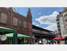 Backsteinfassade eines historischen Bahnhofsgebäudes mit dekorativen Elementen und Rundfenstern. Daneben eine erhöhte Plattform mit Gleisen, flankiert von modernen Wohnhäusern und Straßencafés mit Sonnenschirmen im Vordergrund.