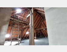 Holzbalkendachboden eines Gebäudes mit sichtbarem offenen Sparrendach, Holzstreben und Ziegeldeckung. Tageslicht fällt durch kleine Dachfenster ein, und eine einfache Holzleiter steht im Vordergrund.