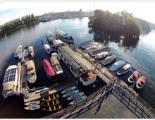 Hafenbereich mit mehreren Booten und kleinen Yachten, die an Stegen vertäut sind. Im Hintergrund ist eine bewaldete Insel sowie eine ruhige Wasserfläche mit Uferbäumen und Gebäuden sichtbar.