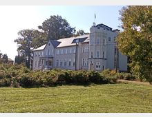 Großes, zweistöckiges Gebäude mit grauer Fassade, zahlreichen Fenstern und einem kleinen Turm auf der rechten Seite. Das Gebäude befindet sich in einer grünen, parkähnlichen Umgebung mit Bäumen und Sträuchern.