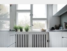 Helle Küche mit großen Fenstern, durch die Tageslicht einfällt. Weiße Schränke und Heizkörperverkleidungen dominieren den Raum, mit grünen Topfpflanzen und einer Obstschale als Dekoration auf der Fensterbank.