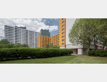 Mehrstöckige Wohngebäude mit weißen und orangefarbenen Fassaden, umgeben von Grünflächen, Bäumen und einem gepflegten Rasen. Im Vordergrund ist eine Hecke zu sehen, der Himmel ist bewölkt.