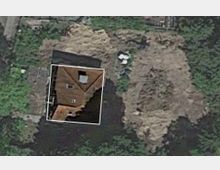 Luftaufnahme eines bewaldeten Bereichs mit einer Baugrube, umgeben von Bäumen. In der Mitte ist ein teilweise freigelegtes Haus mit braunem Dach und der Nummer „28“ markiert.