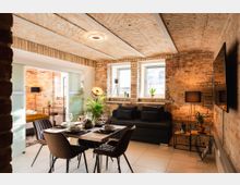 Wohnraum mit industriellem Stil, gekennzeichnet durch freiliegende Ziegelwände und ein abgerundetes Ziegelgewölbe an der Decke. Der Raum ist möbliert mit einem Esstisch, Stühlen, einer schwarzen Couch und dekorativen Pflanzen; im Hintergrund ist ein Schlafbereich durch eine Glas-Schiebetür abgetrennt.