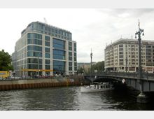 Moderne Bürogebäude mit großen Glasfronten an einem Flussufer, neben einer dekorierten Stahlbrücke mit Laternen. Ein Ausflugsboot fährt auf dem Fluss unter der Brücke hindurch, umgeben von Stadtarchitektur im Hintergrund.