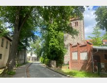Gepflasterte Straße mit Bäumen, die Schatten spenden, flankiert von älteren Gebäuden; rechts ein historischer Feldsteinturm mit Ziegelverzierungen, vermutlich Teil einer Kirche, und ein Ziegelbau im Vordergrund. Der Hintergrund zeigt weitere kleinere Häuser in einem ruhigen, ländlichen Umfeld.