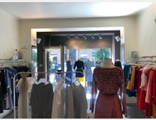 Innenraum eines modernen Bekleidungsgeschäfts mit Kleiderstangen voller Kleidung auf beiden Seiten und Schaufensterpuppen in der Mitte. Im Hintergrund befindet sich ein großes Schaufenster mit Blick auf eine Straße.