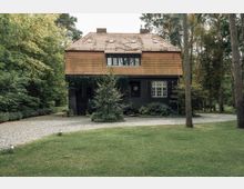 Zweistöckiges Haus mit einem Satteldach aus Ziegeln, einer schwarzen Fassade und weiß gerahmten Fenstern, umgeben von dichtem Baumbestand und gepflegtem Rasen. Ein gepflasterter Weg führt durch den Vorgarten.