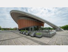 Gebäude mit futuristischem Design und geschwungener Dachkonstruktion, umgeben von einer gepflasterten Fläche und bepflanzten Holzkästen; im Hintergrund sind Bäume und weitere Gebäude erkennbar.