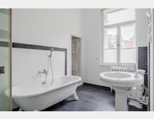 Helles Badezimmer mit freistehender Badewanne an einer weiß gefliesten Wand mit dunkler Mosaikbordüre, großem Fenster und dunkelgrauen Bodenfliesen. Im Vordergrund stehen ein rundes Waschbecken auf Säule und ein Heizkörper unter dem Fenster.