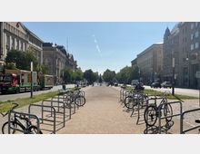 Breiter Fußweg mit Fahrradständern und Bänken in der Mitte, flankiert von historischen Gebäuden und Bäumen. Im Hintergrund ist ein Denkmal erkennbar, umgeben von einer Allee mit Straßenverkehr bei klarem Himmel.