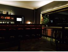 Dunkle Bar mit moderner Einrichtung, einem langen Tresen mit barhockerbestückter Theke, darüber eine beleuchtete Regalwand mit Getränkeflaschen. Große Fensterfront mit Pflanzen in der Ecke; die Beleuchtung ist gedämpft.