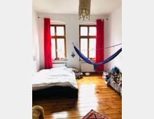 Ein helles Schlafzimmer mit Holzdielenboden, zwei großen Fenstern mit roten Vorhängen und einer blauen Hängematte, die zwischen den Wänden aufgehängt ist. Links steht ein Doppelbett mit weißer Decke, rechts befinden sich Regale und Körbe mit Stofftieren.