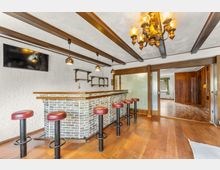 Innenraum mit gemauerter Theke im Barstil, Holzfußboden und mehreren Barhockern mit roten Polstern. Sichtbare Deckenbalken, hängende Leuchten und eine große Glasschiebetür führen in einen angrenzenden Raum mit Holzvertäfelung.