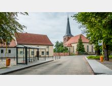 Straßenszene in einer ländlichen Umgebung mit einer Bushaltestelle, einem Wartehäuschen aus Glas und einer gepflasterten Fahrbahn. Im Hintergrund steht eine Kirche mit einem spitzen Turm neben Gebäuden mit roten Ziegeldächern, umgeben von Bäumen und Grünanlagen.