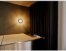 Minimalistischer Innenraum mit einer gepolsterten Liegefläche auf einem Metallpodest, beleuchtet von einer runden Wandleuchte an einer dunkel getönten Wand. Rechts hängen schwere, bodenlange Vorhänge als Raumteiler neben einer hellen Seitenwand.