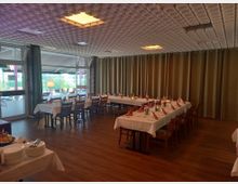 Ein gedeckter Veranstaltungsraum mit zwei langen Tafeln, die weiße Tischdecken, Stühle aus Holz und dekorative Servietten aufweisen. Im Hintergrund befinden sich bodentiefe Fenster mit Vorhängen und eine beige Wand mit Vorhängen, der Boden besteht aus Holzoptik.