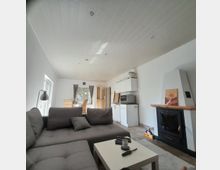 Wohnzimmer mit offener Küche in einem modernen, hellen Design. Im Vordergrund steht eine graue Eckcouch mit einem kleinen weißen Couchtisch, im Hintergrund sind ein Esstisch mit Stühlen, eine einfache Küchenzeile und ein Kaminofen mit dekorativem Bild sichtbar.