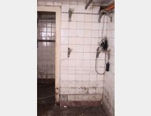 Ein heruntergekommener Raum mit weißen, schmutzigen Fliesenwänden, sichtbaren Halterungen und einem schwarzen, beschädigten Bereich um eine Steckdose. Der Boden ist uneben und verschmutzt, und eine geöffnete Tür führt zu einem weiteren gefliesten Raum.