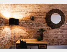 Ecke eines Raumes mit einer Wand aus roten Backsteinen, einem kleinen Holztisch mit schwarzem Metallgestell, einer schwarzen Stuhllehne, einer Stehlampe mit schwarzem Schirm und einem runden Spiegel mit dekorativem Holzrahmen. Auf dem Tisch stehen zwei Pflanzen, darunter ein kleiner Topf mit grünem Blattwerk.