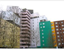 Mehrstöckige Wohngebäude mit farbenfrohen Fassaden in Weiß, Gelb, Grün, Braun und Rosa. Vor den Gebäuden stehen kahle Bäume, und der Himmel ist bewölkt.