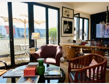 Wohnzimmer mit großen, bodentiefen Fenstern, die auf einen Balkon führen. Der Raum ist mit einem Sessel, einem Glastisch, dekorativen Accessoires und einem Schreibtisch mit Büchern und Kunstobjekten eingerichtet; auf dem Balkon stehen ein Tisch mit Stühlen und Sonnenschirme.