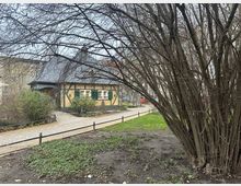 Ein kleiner Fachwerkbau mit gelber Fassade, grünen Fensterläden und dunklem Schieferdach, umgeben von Gehwegen, Sträuchern und einem Baum im Vordergrund, in einer parkähnlichen Umgebung.