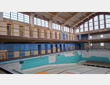 Ein leerstehendes Hallenbad mit abgeblättertem Putz und Farbresten an den Wänden. Die Hauptebene zeigt eine leere Schwimmhalle mit türkisfarbenen Kacheln, umgeben von blauen Umkleidekabinen und einer Empore mit Geländern unter einer hohen Fensterfront.
