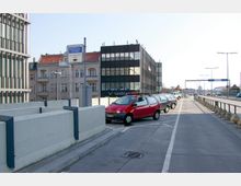 Straßenszene mit Parkplätzen und einem „Parkschein-Automat“-Schild im Vordergrund; im Hintergrund sind ein modernes Bürogebäude mit dunkler Glasfassade, ältere Wohngebäude mit roten Dächern und ein erhöhtes Straßennetz sichtbar. Links und rechts des Weges befinden sich Betonbarrieren und geparkte Fahrzeuge.