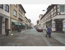 Eine gepflasterte Fußgängerzone in einer Altstadt mit zweistöckigen Gebäuden auf beiden Seiten, teils in Fachwerkbauweise. Einige Geschäfte mit Schaufenstern und Markisen säumen die Straße, während Passanten und ein Hund im Vordergrund zu sehen sind.