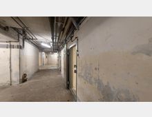 Langer, schmaler, unterirdischer Flur mit abgenutzten, hellen Wänden, sichtbaren Rohrleitungen an der Decke und mehreren schweren Metalltüren. Der Boden besteht aus Beton, und der Flur wird von neonartigen Deckenleuchten erhellt.