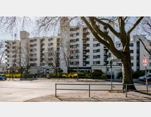 Mehrstöckiges Wohngebäude in heller Farbe mit geometrischen Fensterformen, umgeben von kahlen Bäumen und einem begrünten Straßenrand. Im Vordergrund eine Kreuzung mit Straßenschildern und einem Gehweg.