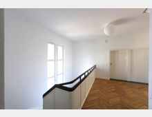 Flur in einem modernen Gebäude mit hellem Holzparkettboden, weiß gestrichenen Wänden und einem schwarzen, an einer Treppe entlang verlaufenden Geländer. Links befindet sich ein großes Fenster, rechts eine weiße Tür.