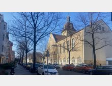 Straßenszene mit Altbaugebäuden, darunter ein großes, helles Gebäude mit Ziegeldach, vielen Fenstern und einem kleinen Turm. Im Vordergrund parken Autos entlang einer Straße mit kahlen Bäumen.