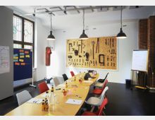 Ein moderner Besprechungsraum mit einem langen Holztisch, umgeben von bunten Stühlen, Notizblöcken und Getränken. Im Hintergrund befinden sich eine Werkzeugwand, ein Boxsack und ein Flipchart; die Raumgestaltung umfasst große Fenster, helle Wände und Industrieleuchten.