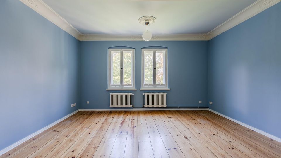 Leerer Wohnraum mit hellblauen Wänden, breiten Holz-Dielenboden und weißer Stuckleiste an der Decke. An der Stirnwand befinden sich zwei hohe Fenster über zwei weißen Heizkörpern, mittig hängt eine runde Deckenleuchte.