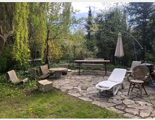 Gartenbereich mit einer gepflasterten Fläche, umgeben von Bäumen und Sträuchern. Auf der Fläche stehen verschiedene Gartenmöbel, darunter Betonstühle, eine weiße Liege, ein runder Tisch, ein Sonnenschirm und ein Grill. Im Hintergrund sind Bänke und dichtes Grün zu sehen.